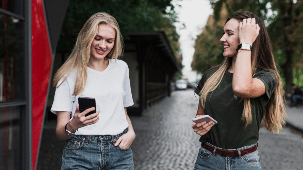 Dwie młode kobiety rozmawiające na ulicy, trzymające telefony, uśmiechnięte – przełamują strach przed mówieniem w obcym języku