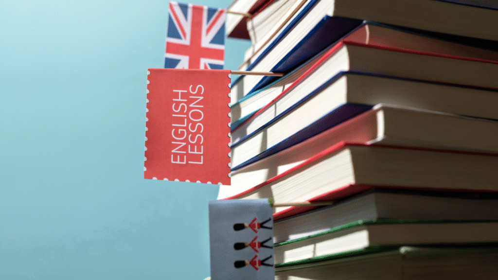 Na obrazku widać stos kolorowych książek, do których przyczepione są małe flagi, w tym brytyjska flaga, czerwona kartka z napisem "ENGLISH LESSONS" oraz obrazek przedstawiający strażników królewskich; całość ilustruje motyw nauki języka angielskiego.